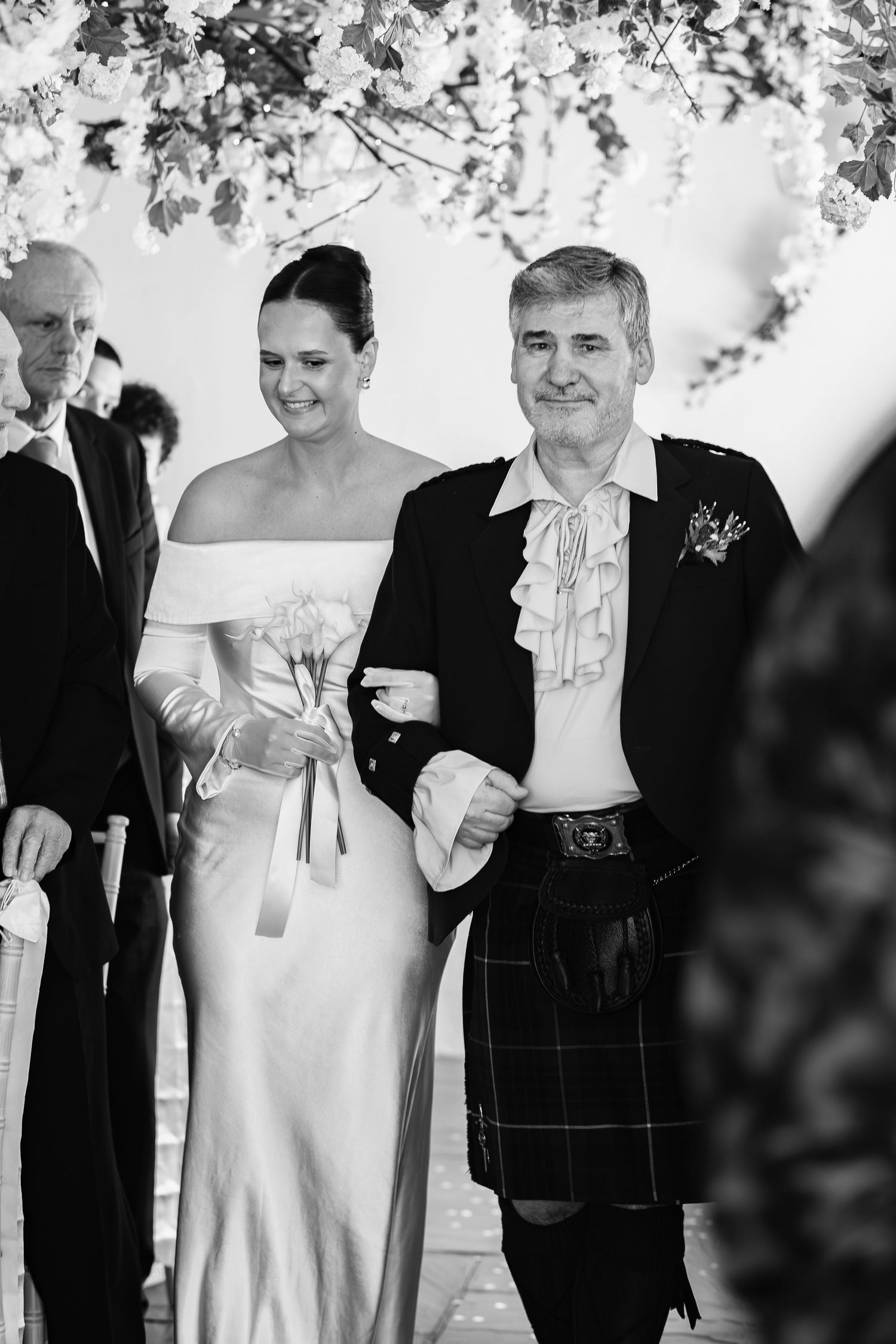 Black and white photo of a bride in a satin gown walking down the aisle with a man in a Scottish kilt.