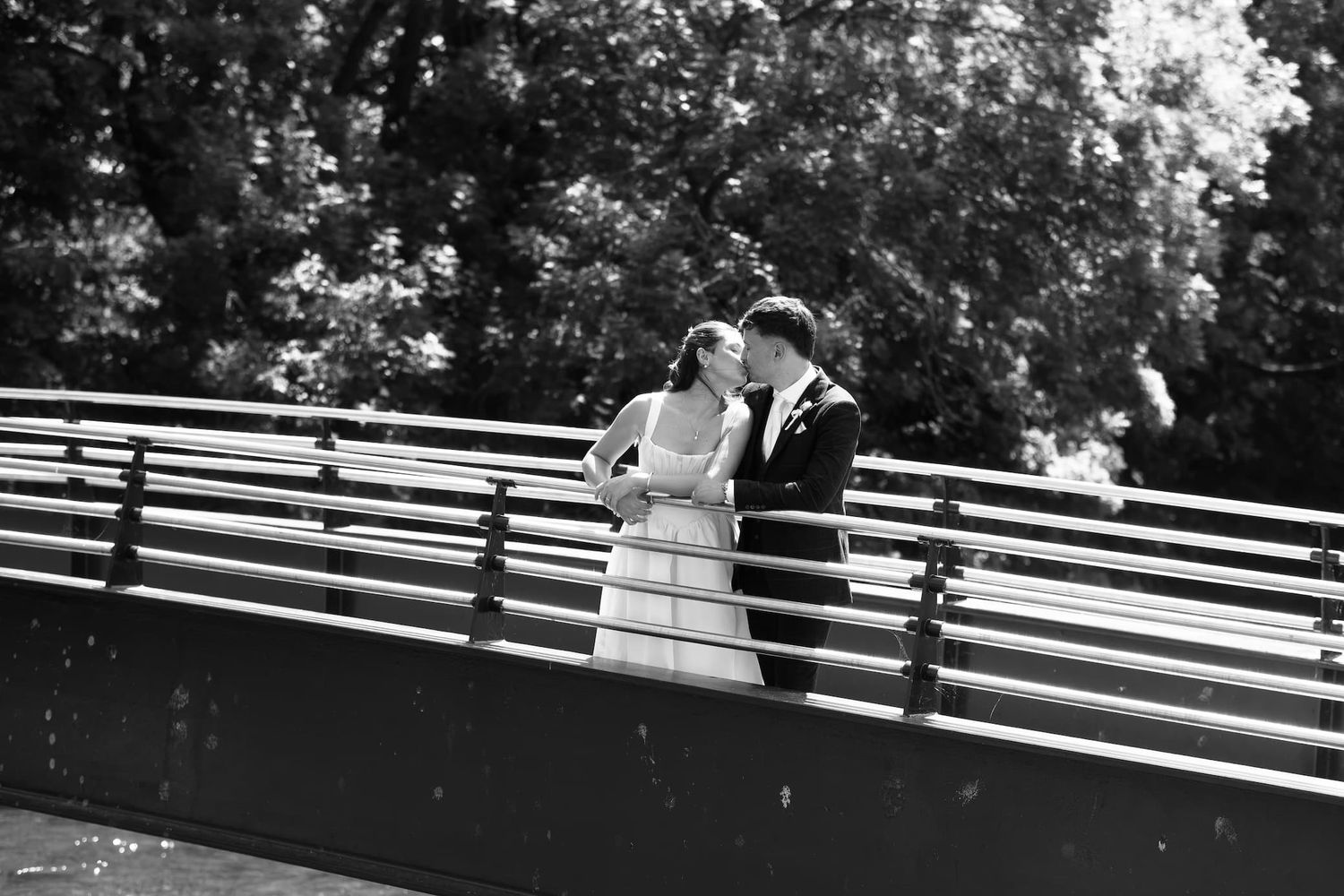 Black and white photo of a bride and groom sharing a romantic moment on a bridge surrounded by lush trees.