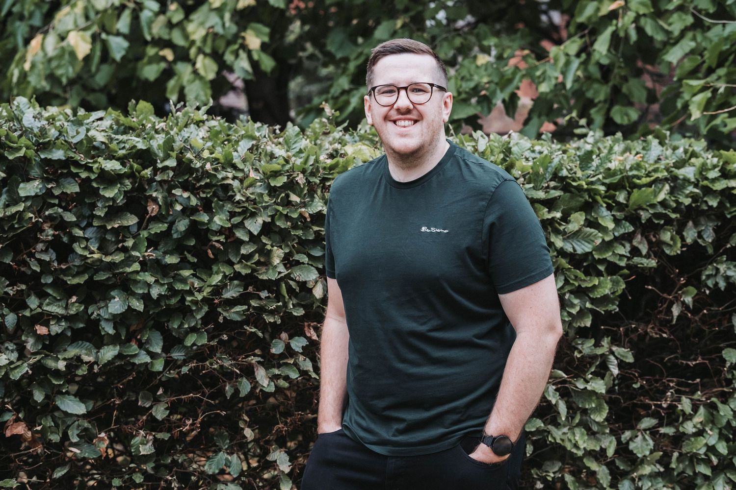 Someone wearing glasses and a dark t-shirt poses outdoors against a green hedge background.
