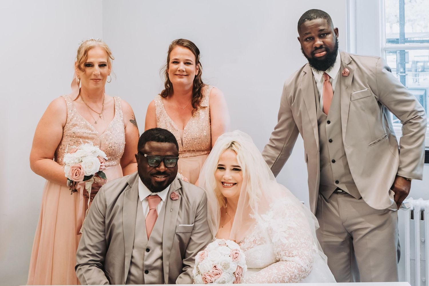 Wedding party members in gray suits and light floral dresses pose together for formal photos.