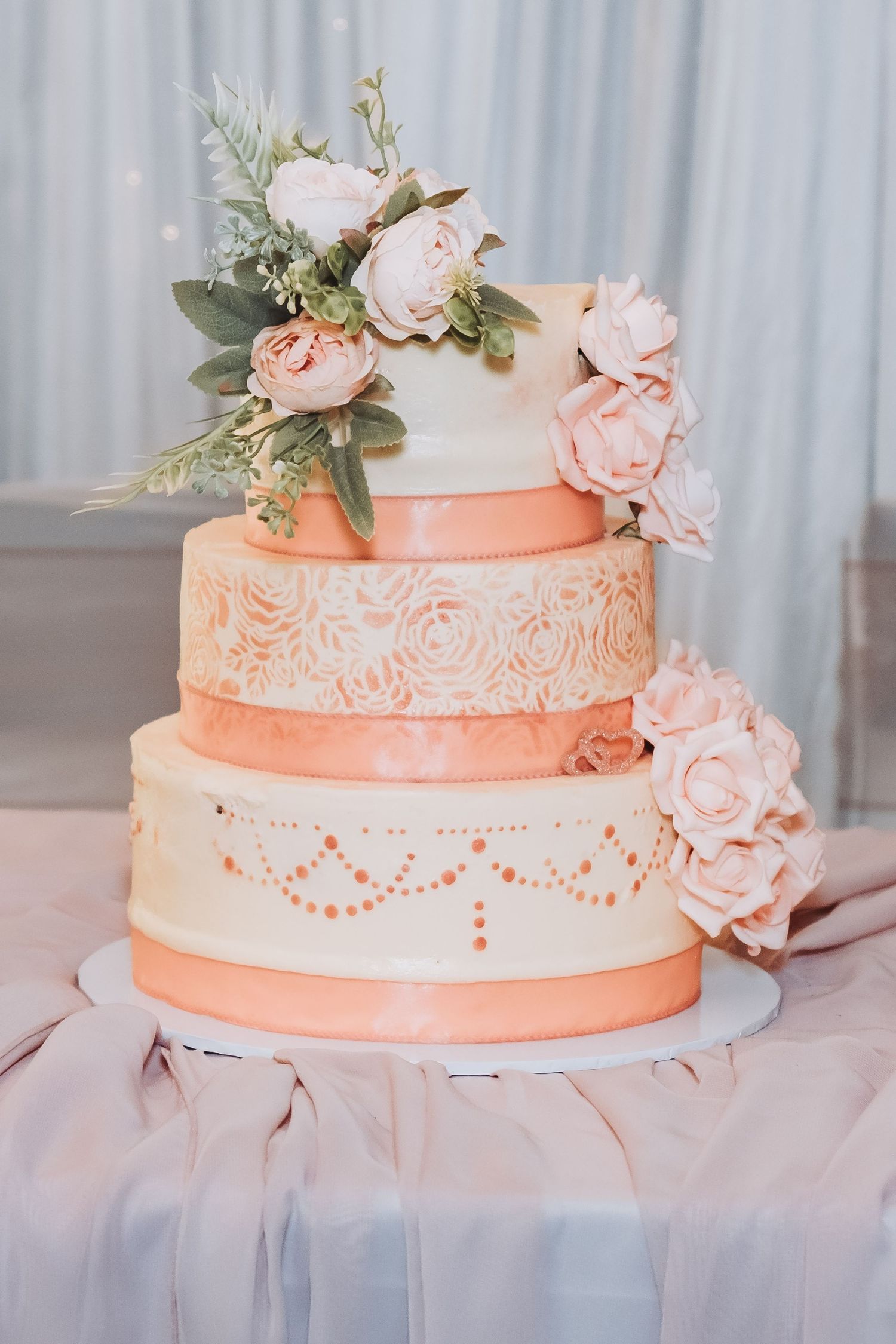 Elegant three-tier peach wedding cake decorated with white lace details and pink roses.