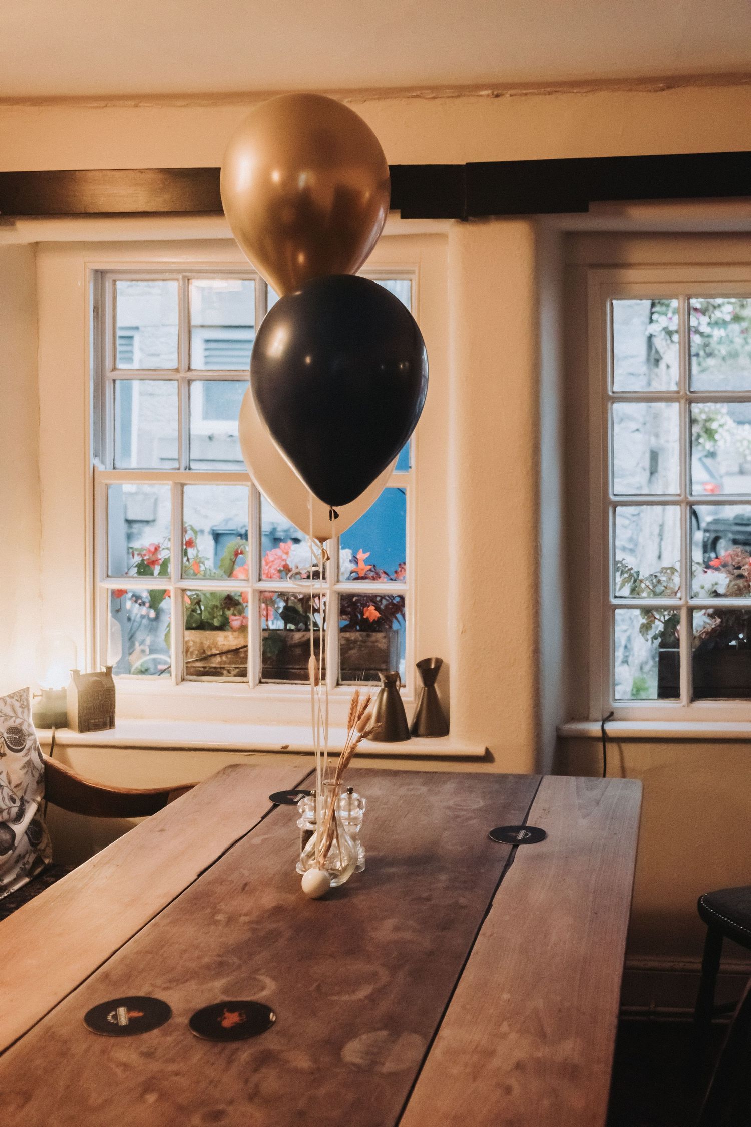 Rustic wooden table setup with large black and gold balloons in an indoor venue with natural lighting through windows.