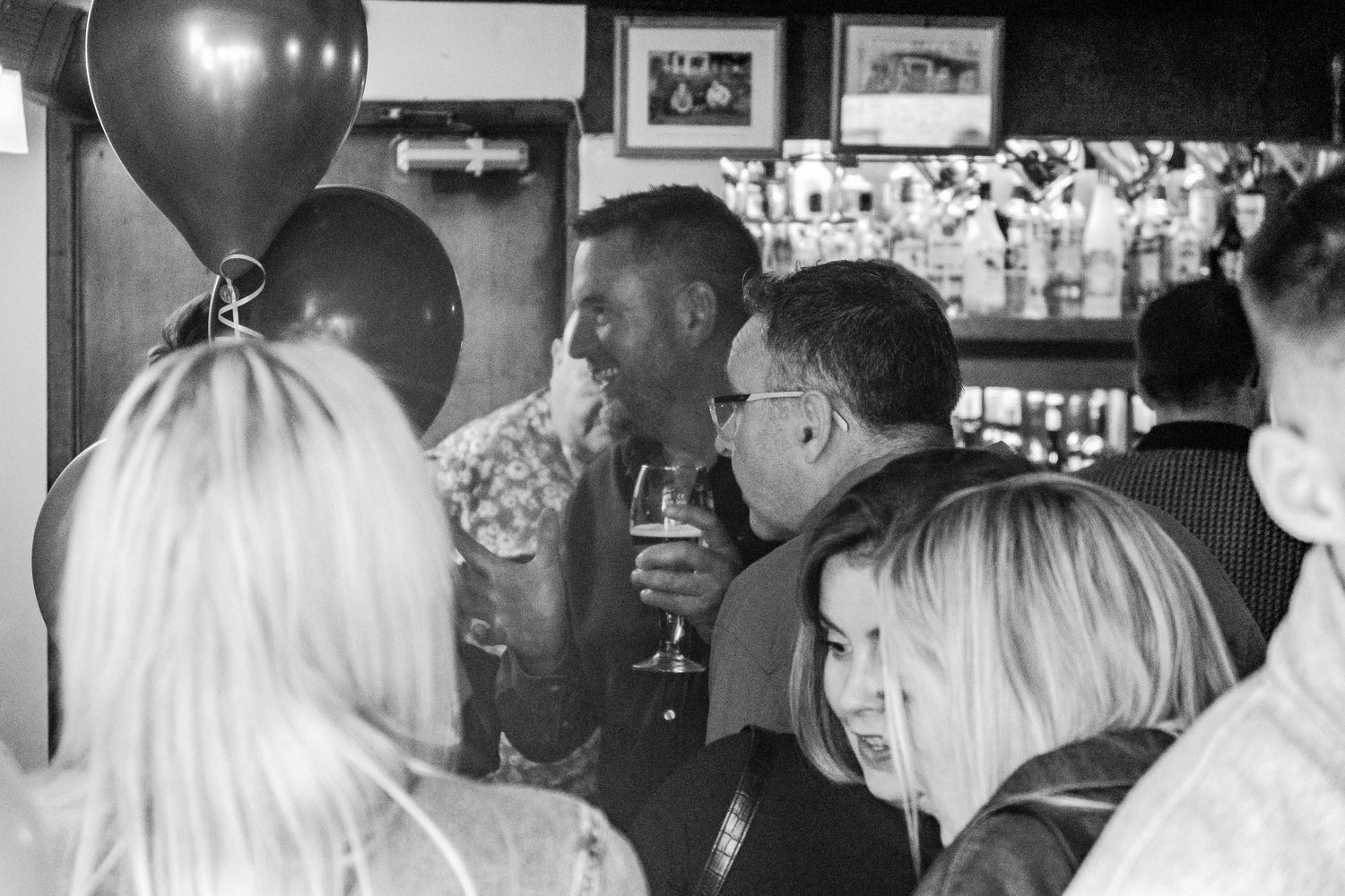 A crowded party scene with balloons and framed photos visible on the walls.