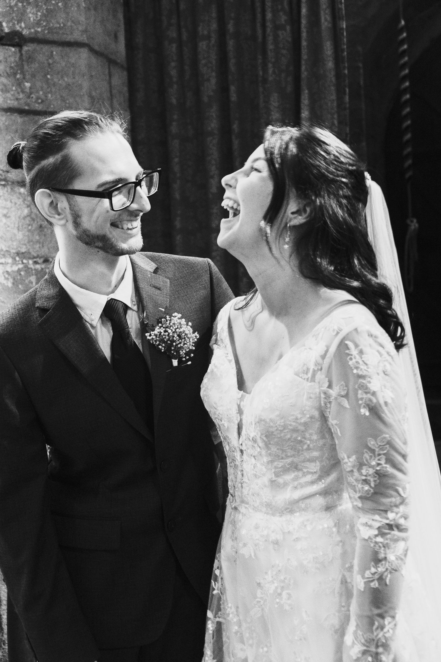 A joyful black and white wedding photo showing a bride laughing while standing next to her groom in a suit and glasses.