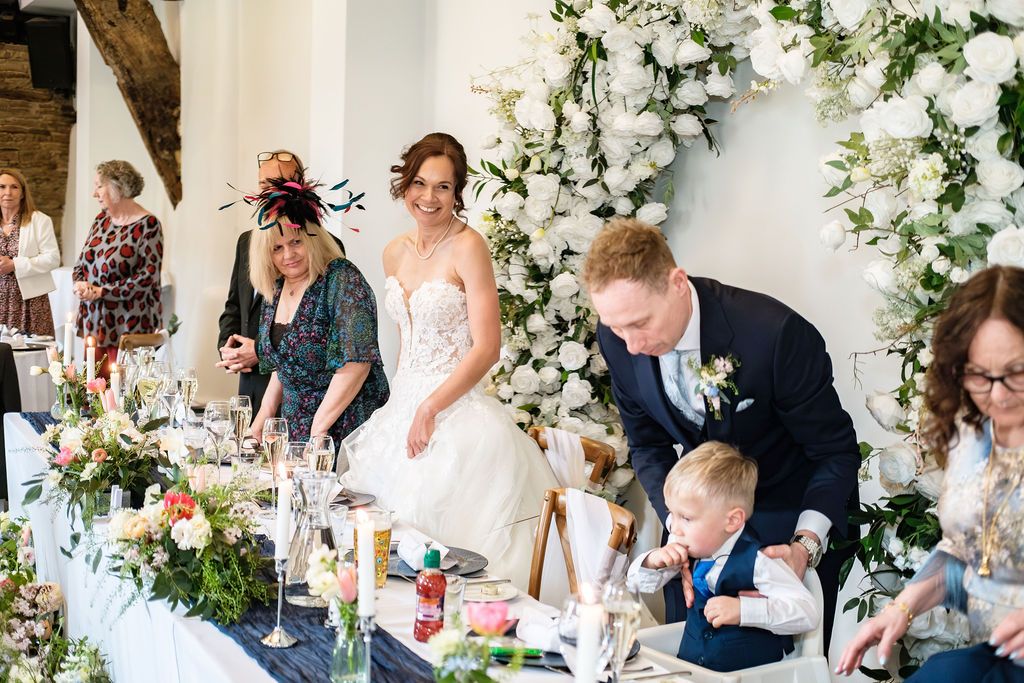 Bride and groom at the top table in front of a white floral arch, child guest nearby