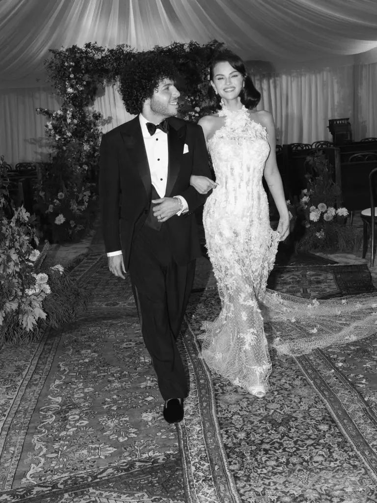 A couple walks arm in arm at a formal event, with the woman wearing an elegant white lace gown and the man in a tuxedo.