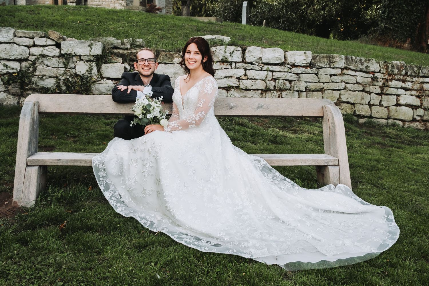 A couple poses for romantic wedding photos on a stone wall backdrop in a garden setting.