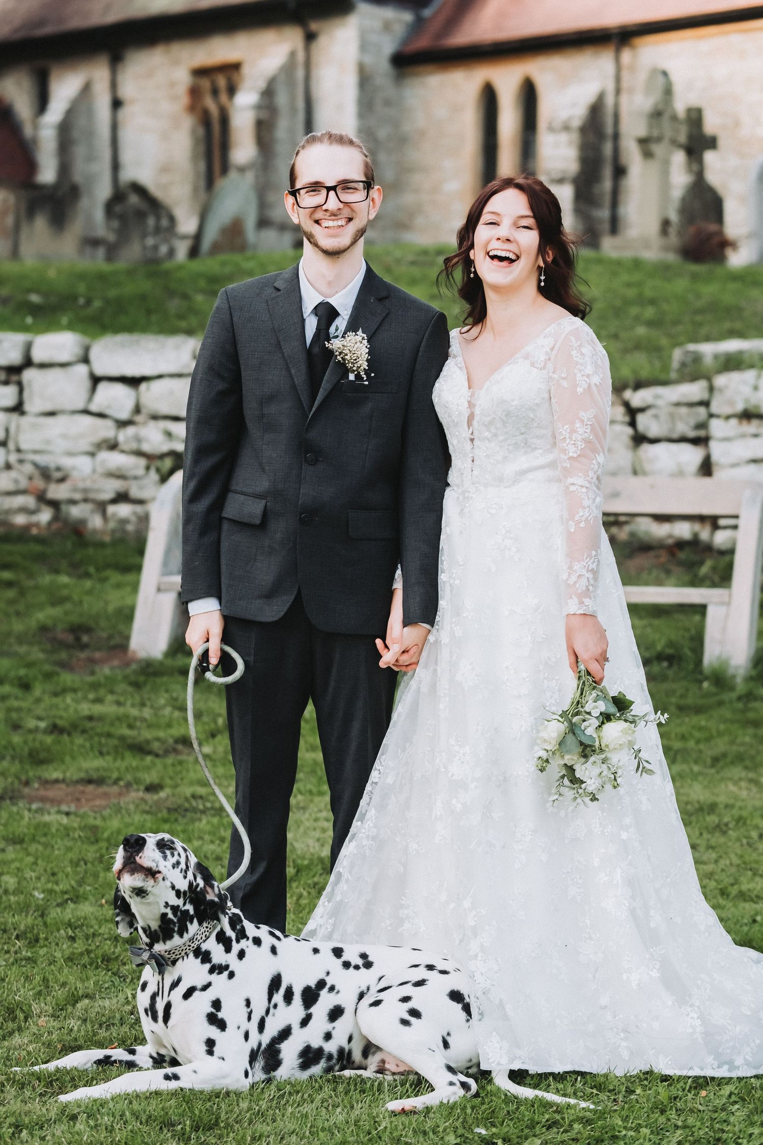 A wedding couple poses with their Dalmatian dog on the church grounds.