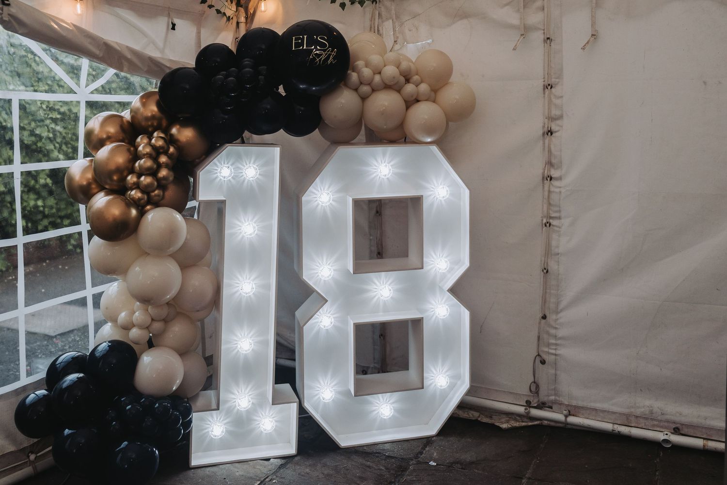 Black and gold balloon arch with illuminated letters creates an elegant party decoration backdrop with luxurious ambiance.
