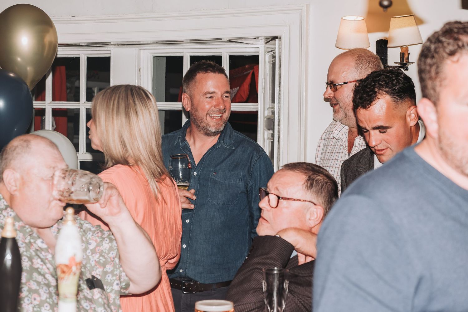 People gathered in groups at an indoor social event with white walls and bright lighting.