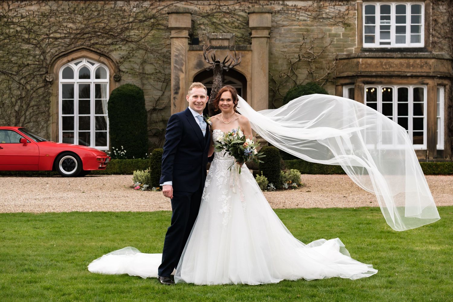 Bride and groom pose on lawn before manor house, her veil billowing in wind, red sports car visible behind them.
