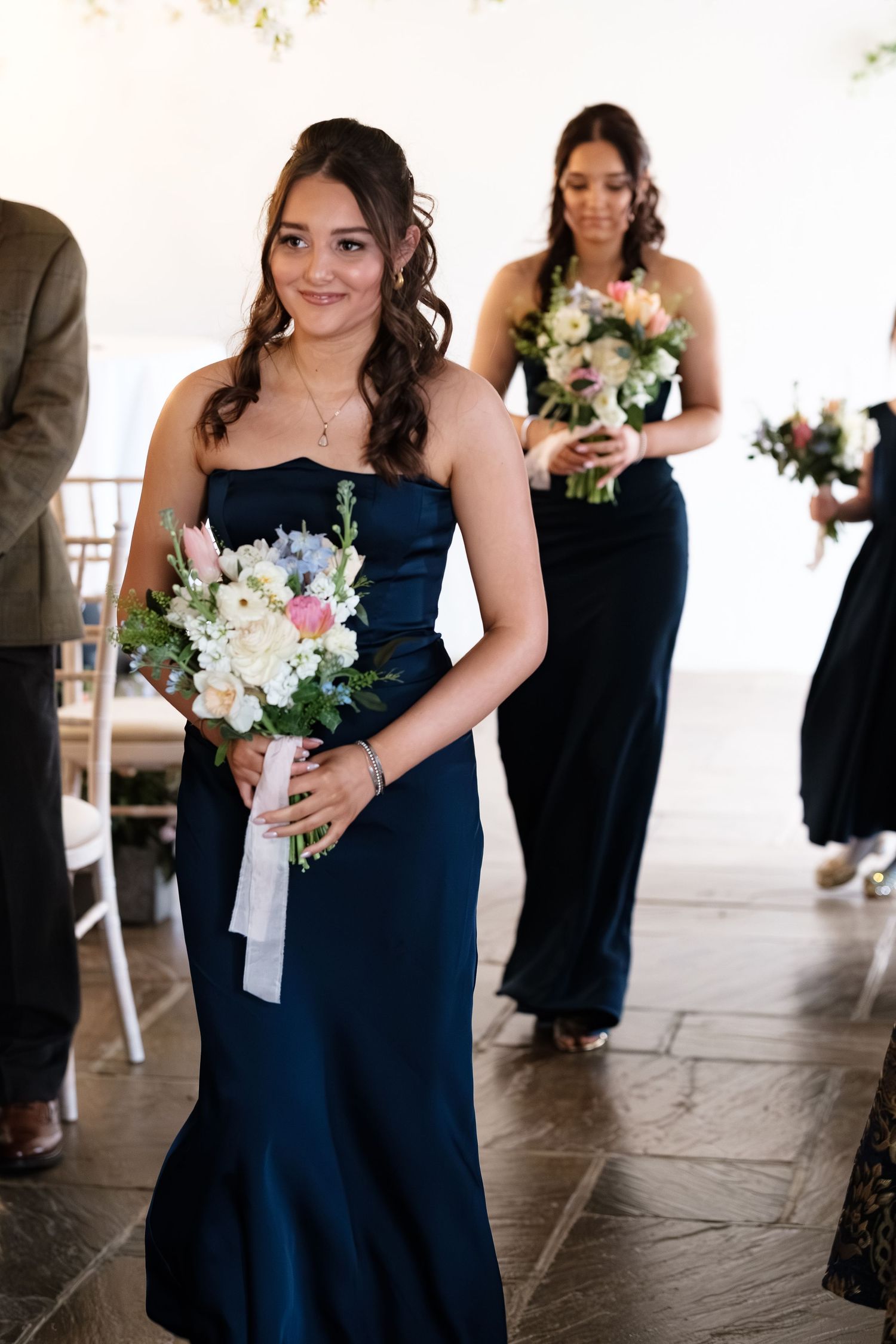 Bridesmaid in navy strapless gown walks down aisle holding white floral bouquet with ribbon at wedding ceremony.