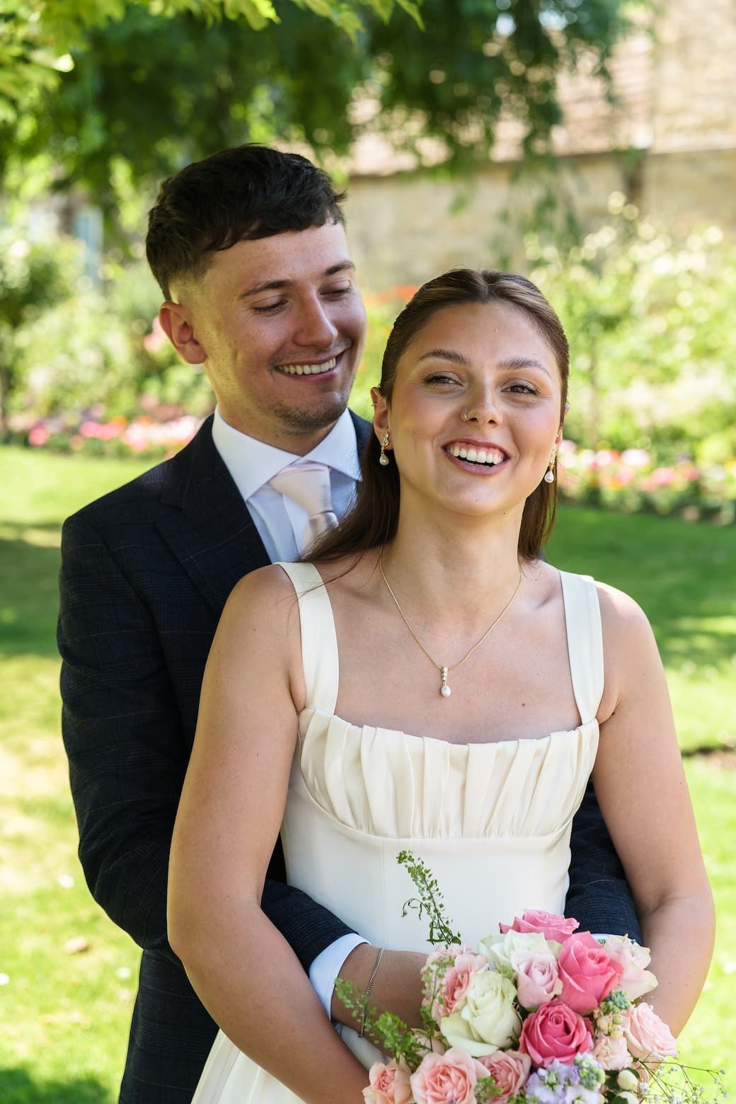 Smiling bride in white dress holding pink bouquet with groom in dark suit embracing her outdoors in garden.