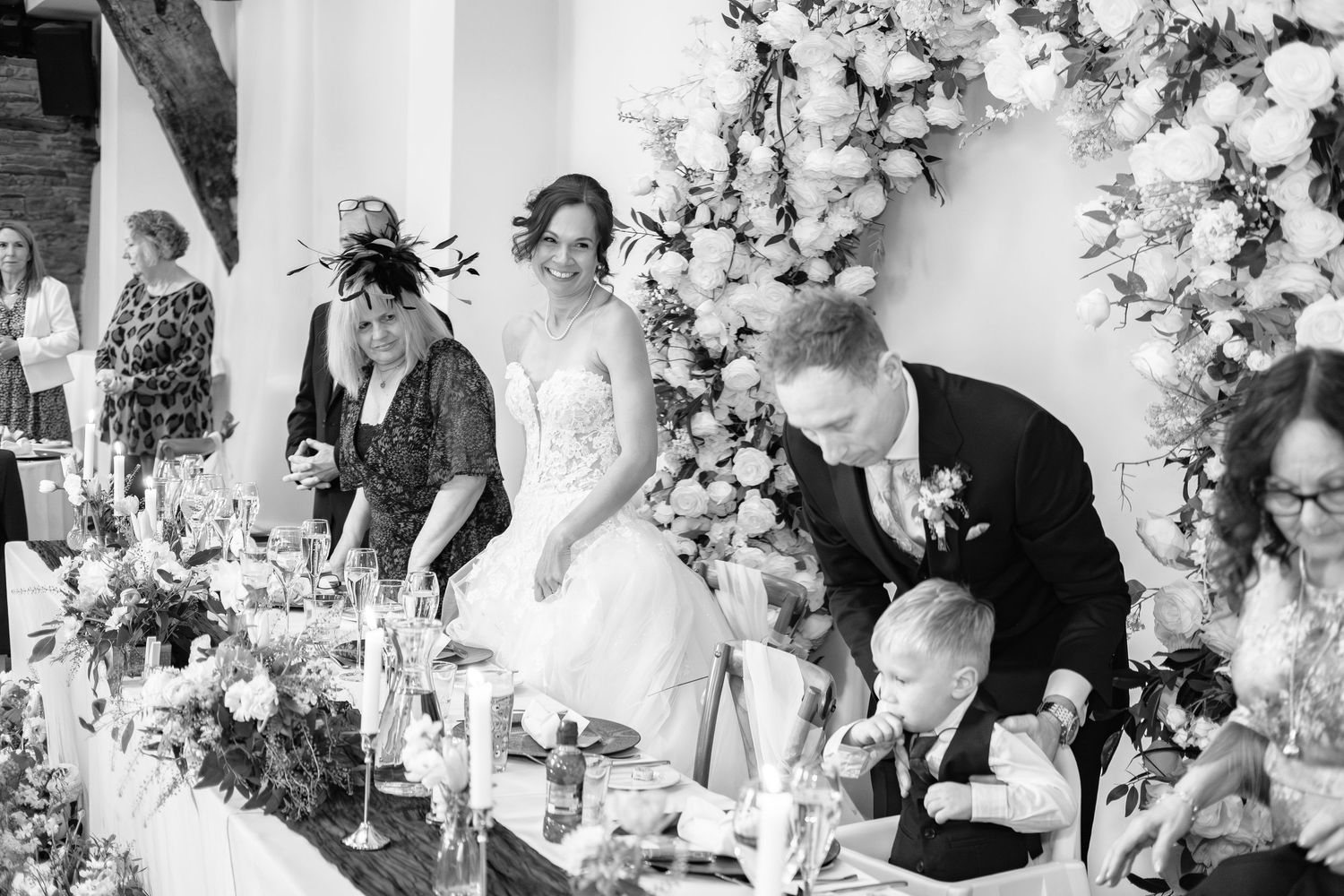 Black and white wedding reception photo showing bride and groom at head table with floral arch backdrop.