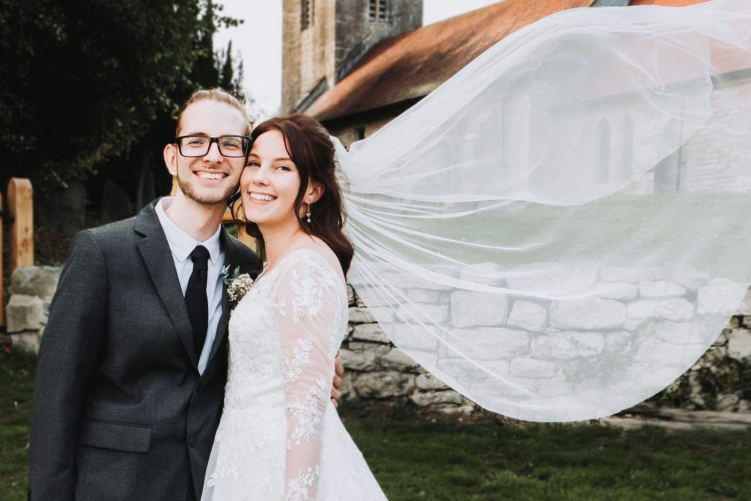 A flowing white veil catches the wind as a couple shares a romantic moment at a rustic stone venue.