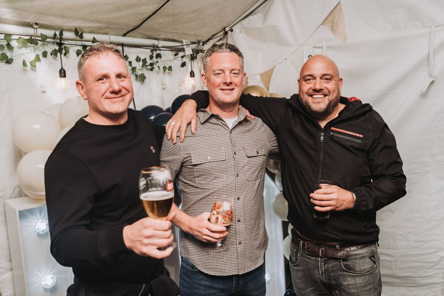 Three friends enjoying drinks together under a white tent at a casual outdoor event.