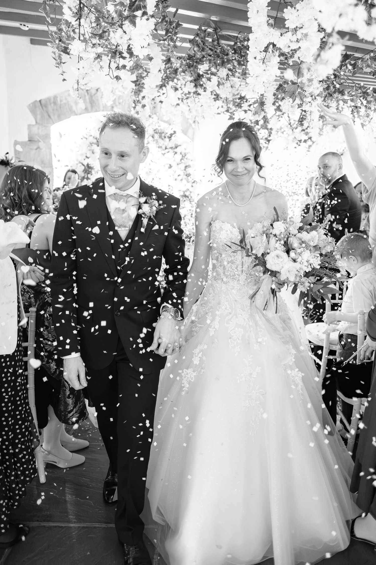 Black and white photo of a newlywed couple walking through confetti shower after wedding ceremony.