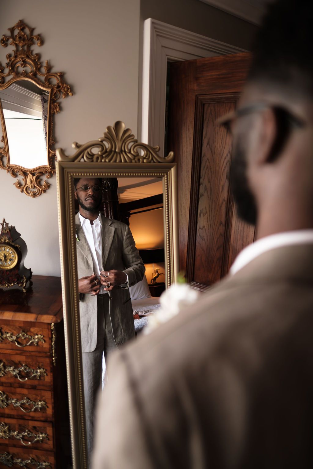 Groom adjusting his jacket in a tall gold-framed mirror
