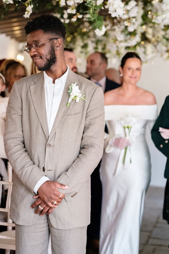 Emotional groom in beige suit watches bride approach in white off-shoulder gown during outdoor wedding ceremony.
