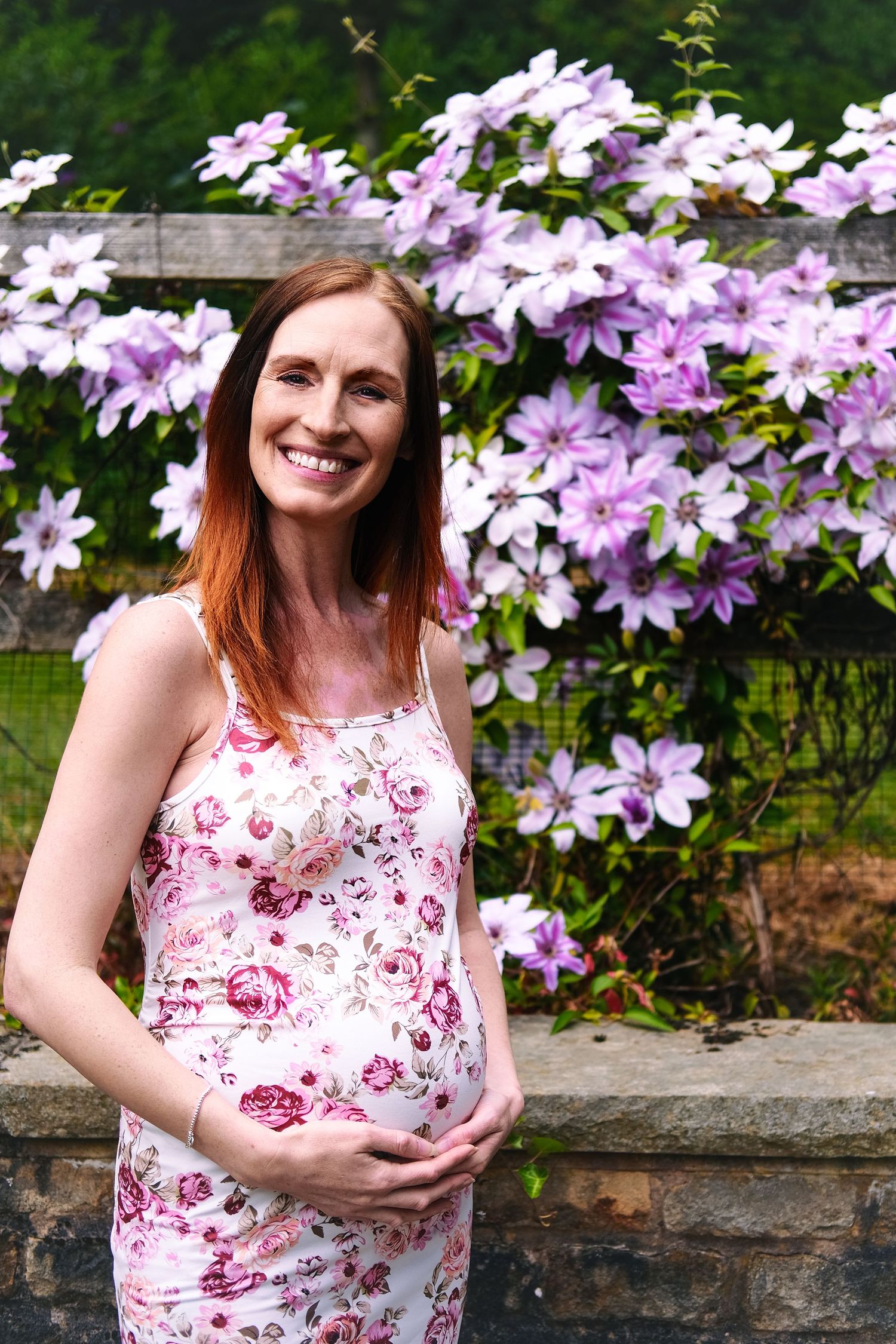 Maternity photos taken against a backdrop of blooming purple clematis flowers on a stone wall.