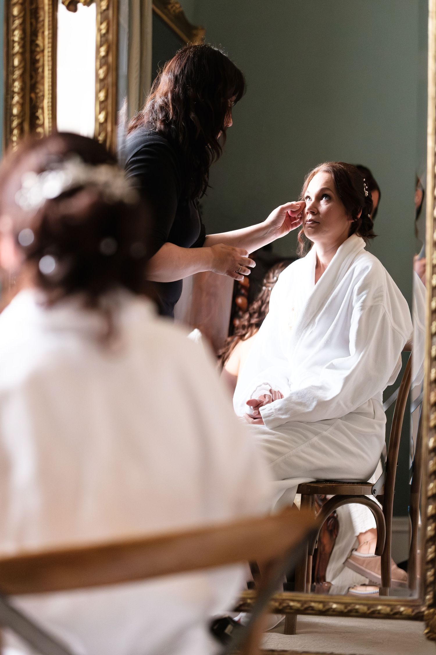 Makeup artist applying finishing touches to a bride in a white robe, seated before an ornate gold mirror.