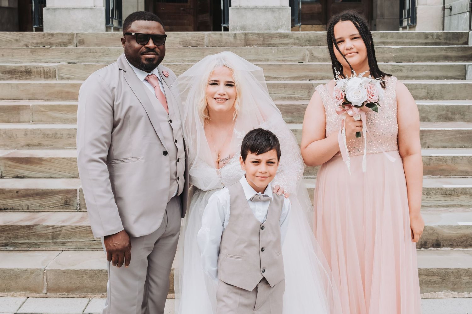 Wedding party in pastel pink dresses and grey suits gather on outdoor stone steps.