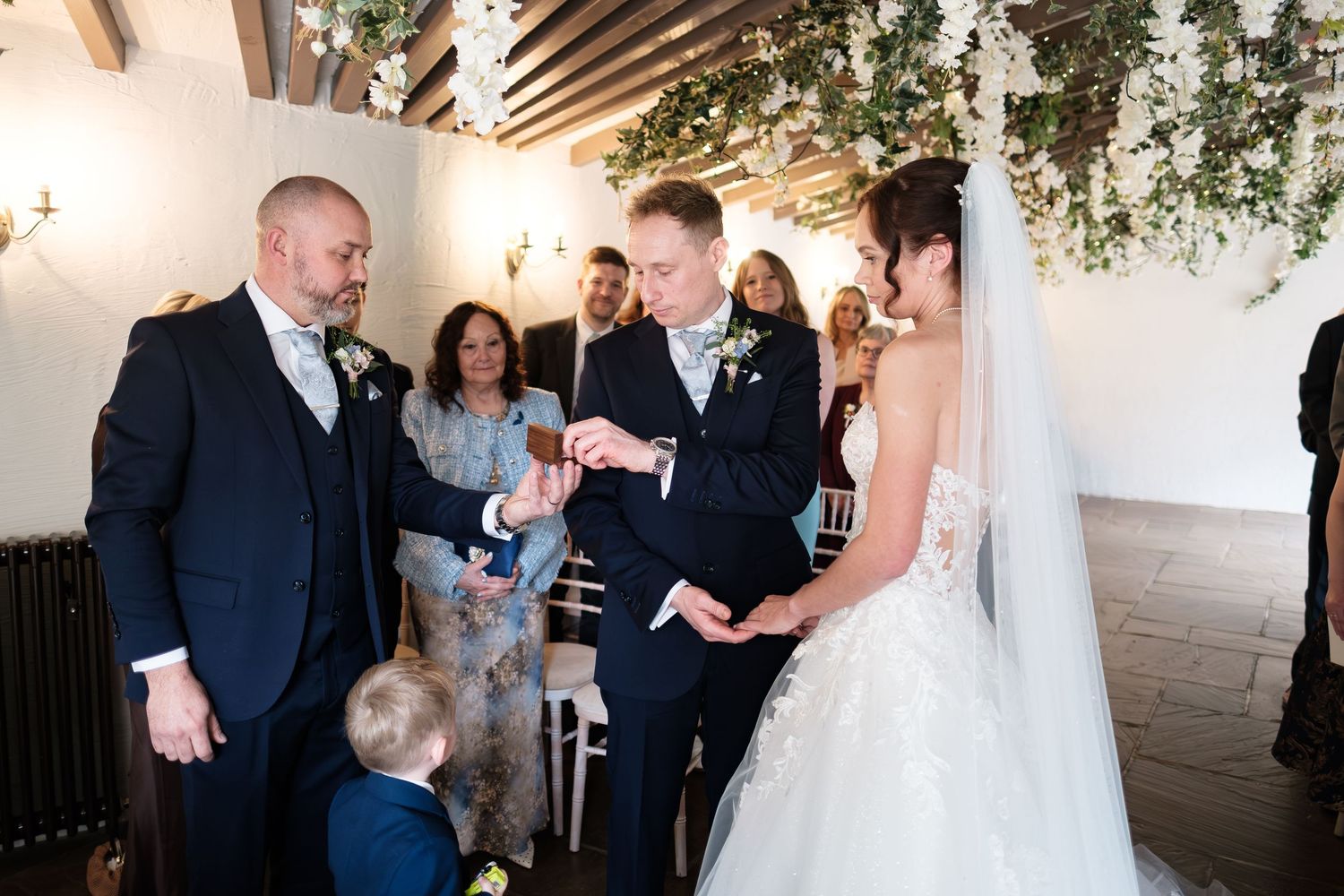 Groom places wedding ring on bride's finger during indoor ceremony with floral arch and guests watching.