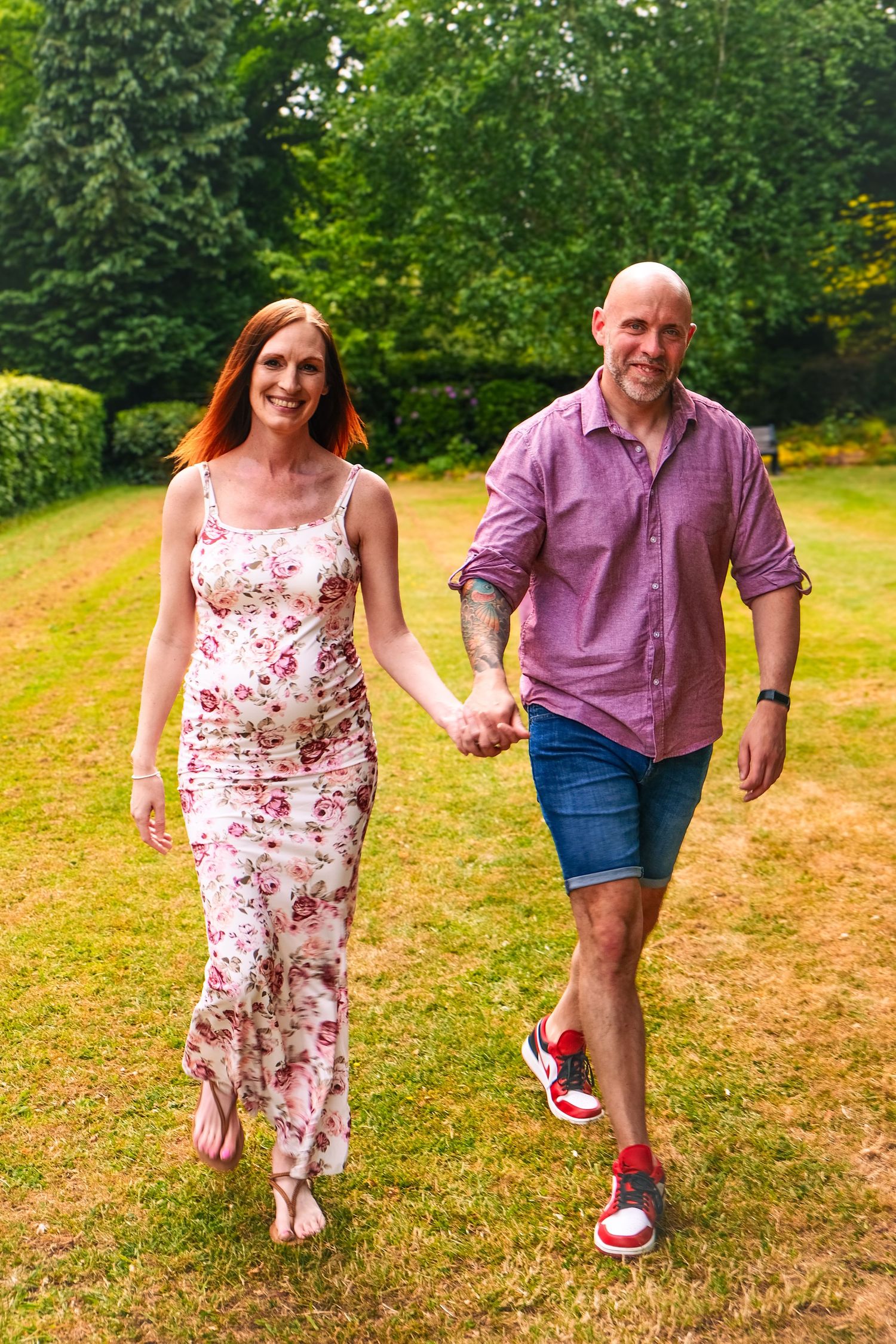 Couple walking outdoors in a grassy field, woman in floral dress and man in purple shirt.