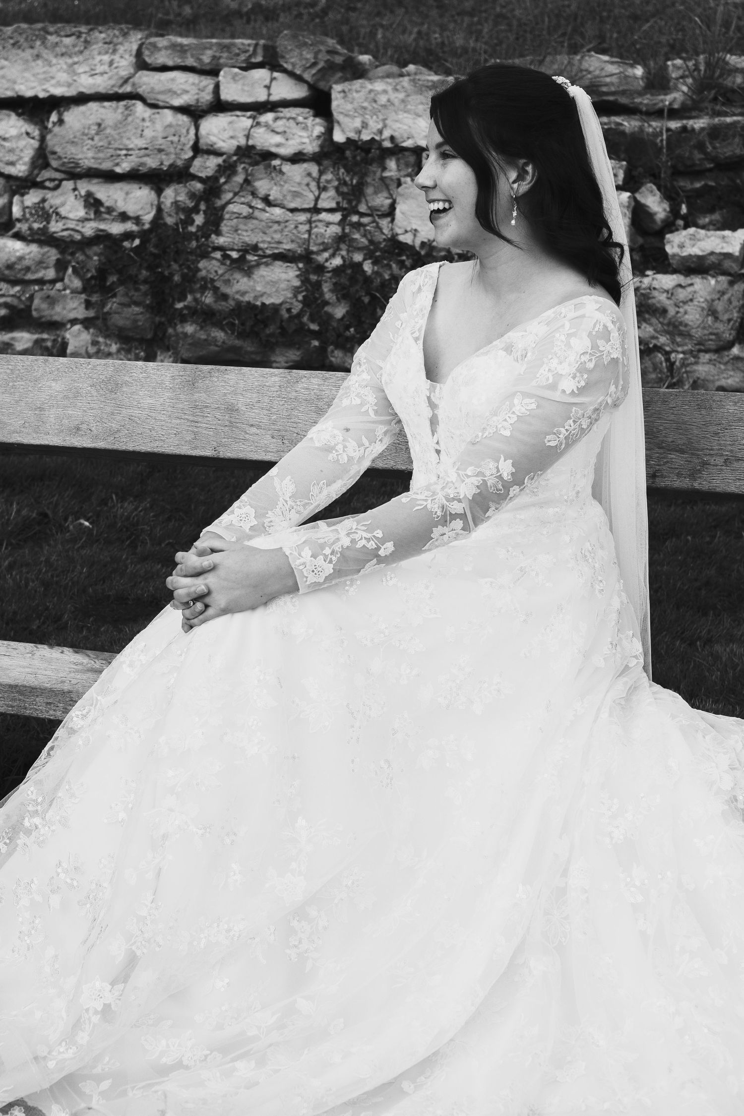 A bride in a flowing white lace wedding gown sits gracefully on stone steps in a black and white photograph.