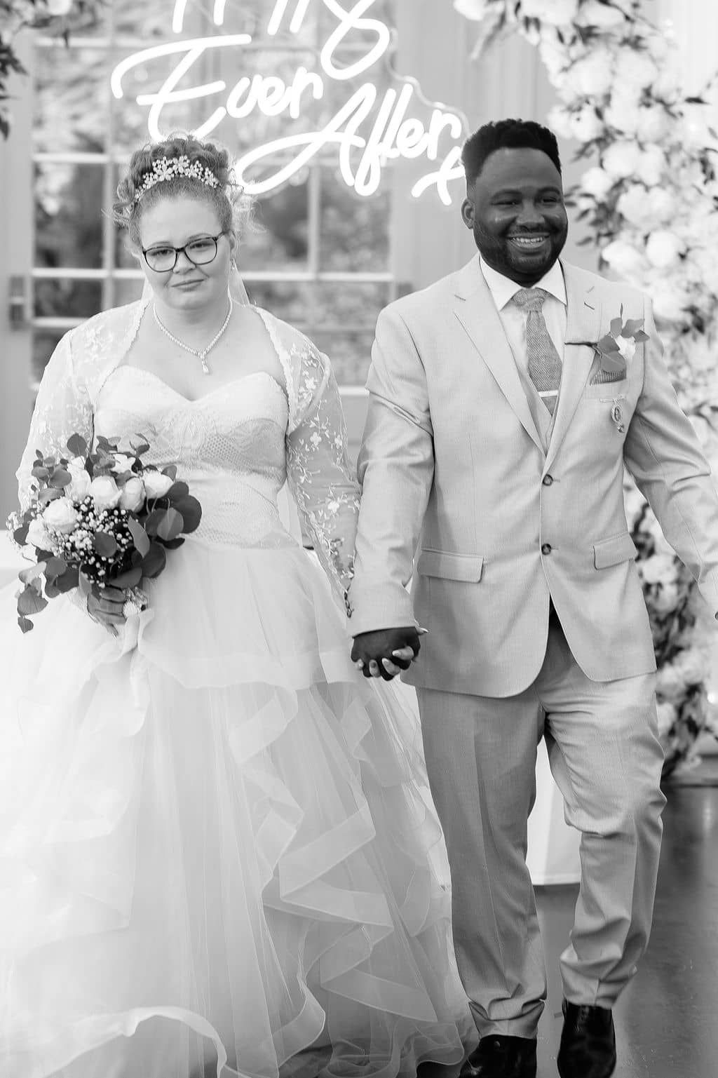 Black and white photo of a smiling bride and groom holding hands at their wedding ceremony altar.