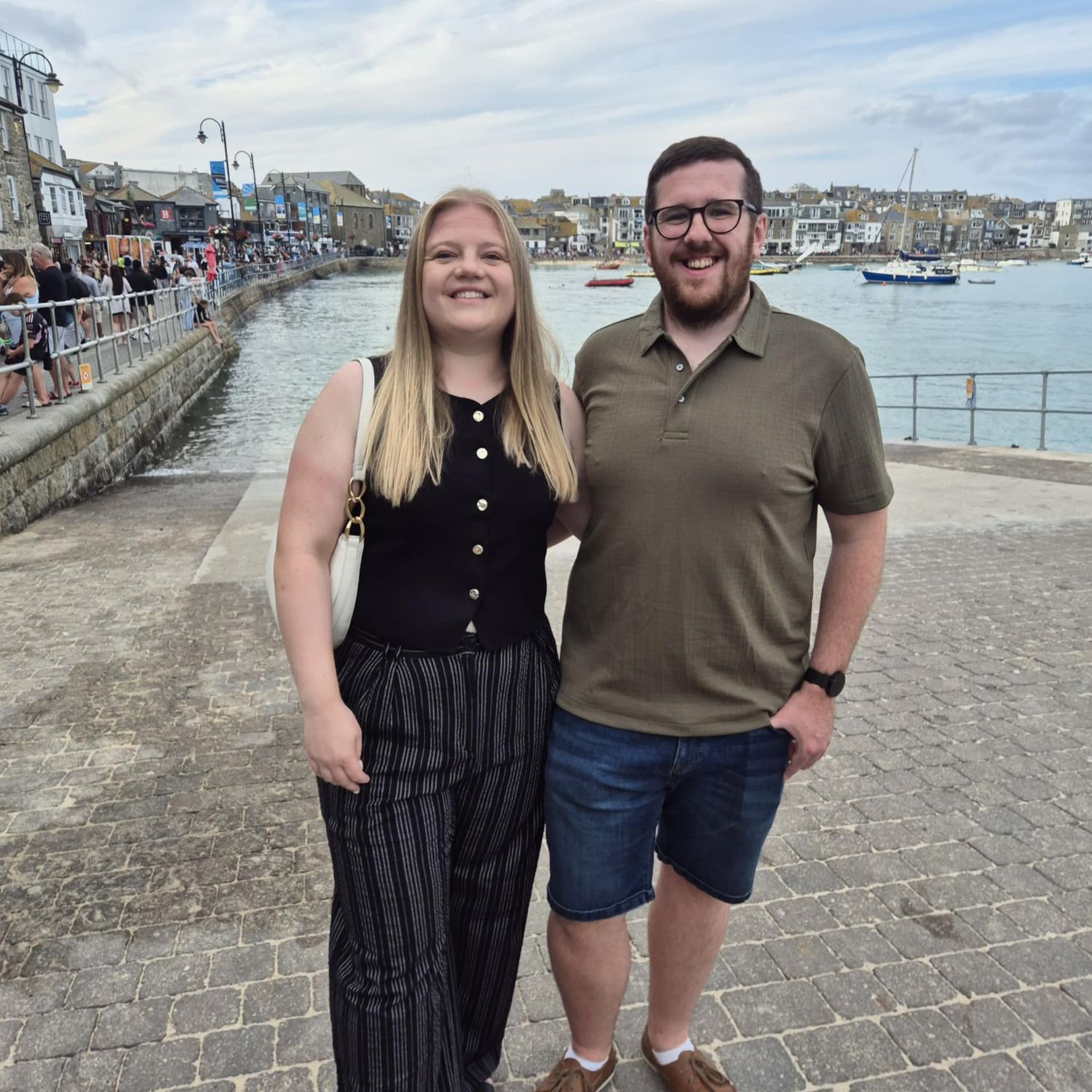 Couple standing on a harbor walkway with boats and waterfront buildings in the background