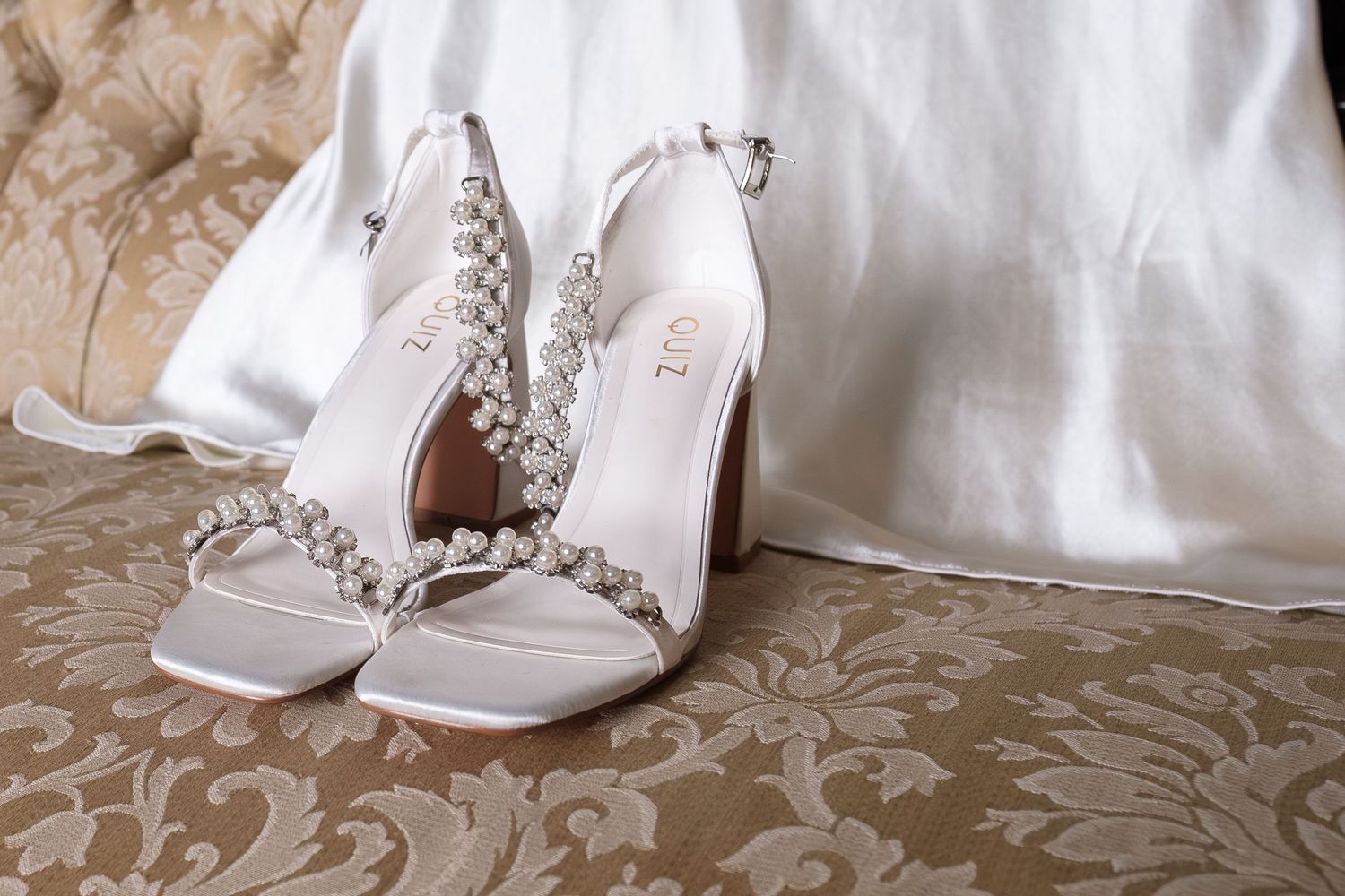 Elegant white bridal heels adorned with pearl and crystal embellishments displayed on gold damask fabric beside a wedding dress.