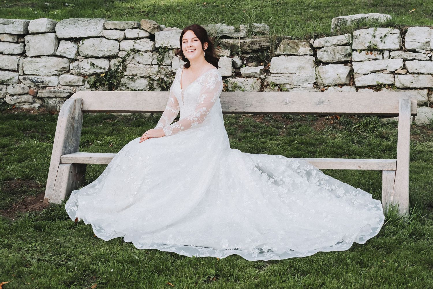 A bride in a flowing white lace gown poses gracefully on a rustic wooden bench against a stone wall backdrop.