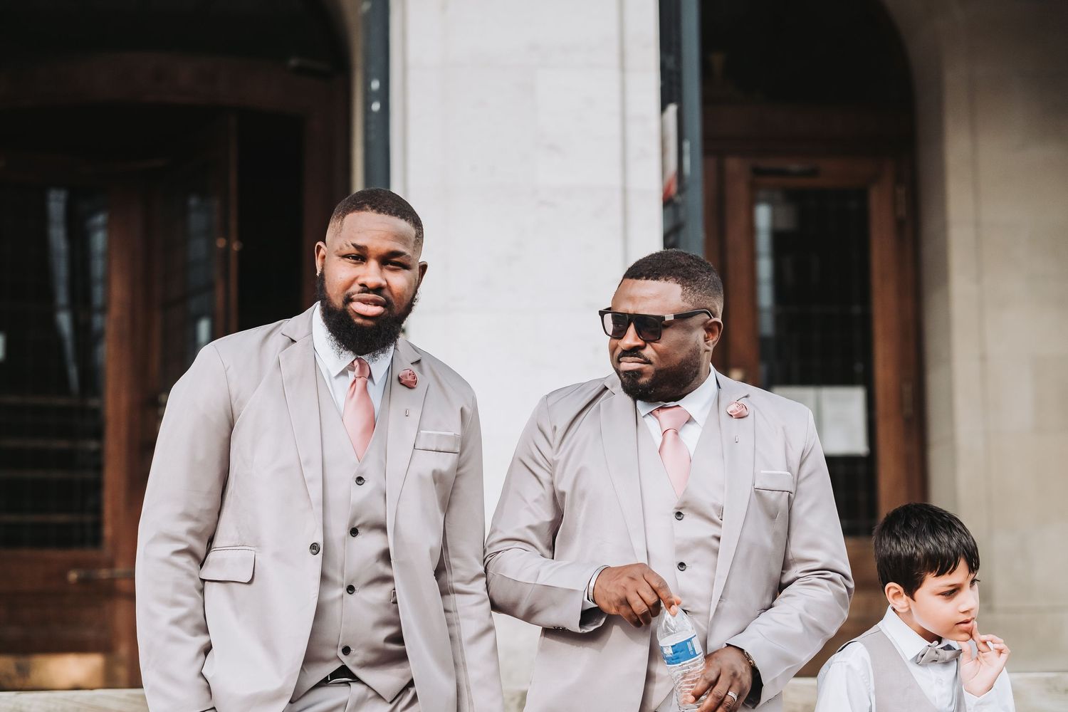 Groomsmen in light grey suits with coral accents stand together outside a building.