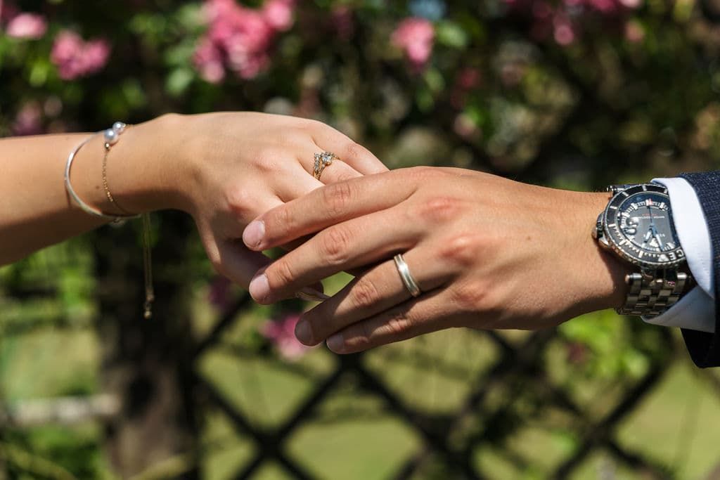 Couple exchanging wedding rings outdoors at Bakewell, bride wearing bracelet and ring, groom wearing a watch, with flowers in background.