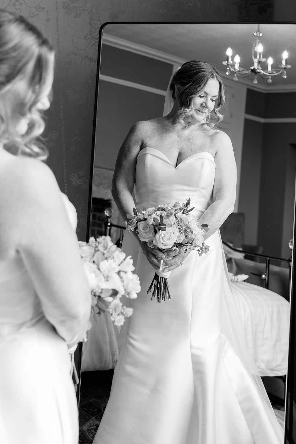 Bride in strapless satin gown holds floral bouquet while admiring her reflection in a large mirror, black and white photo.