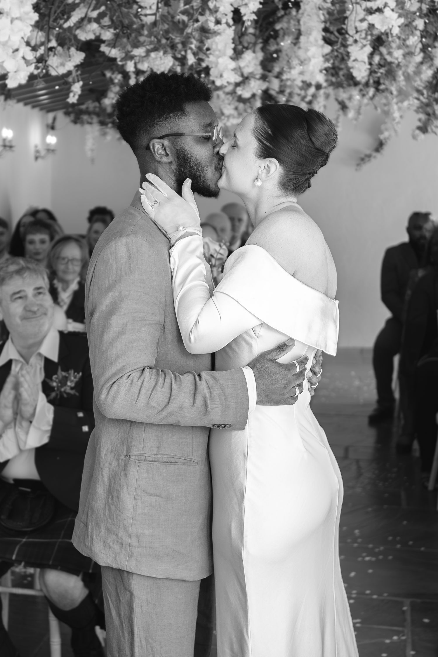 Black and white photo of a couple sharing their first kiss at a wedding ceremony, surrounded by floral decorations.