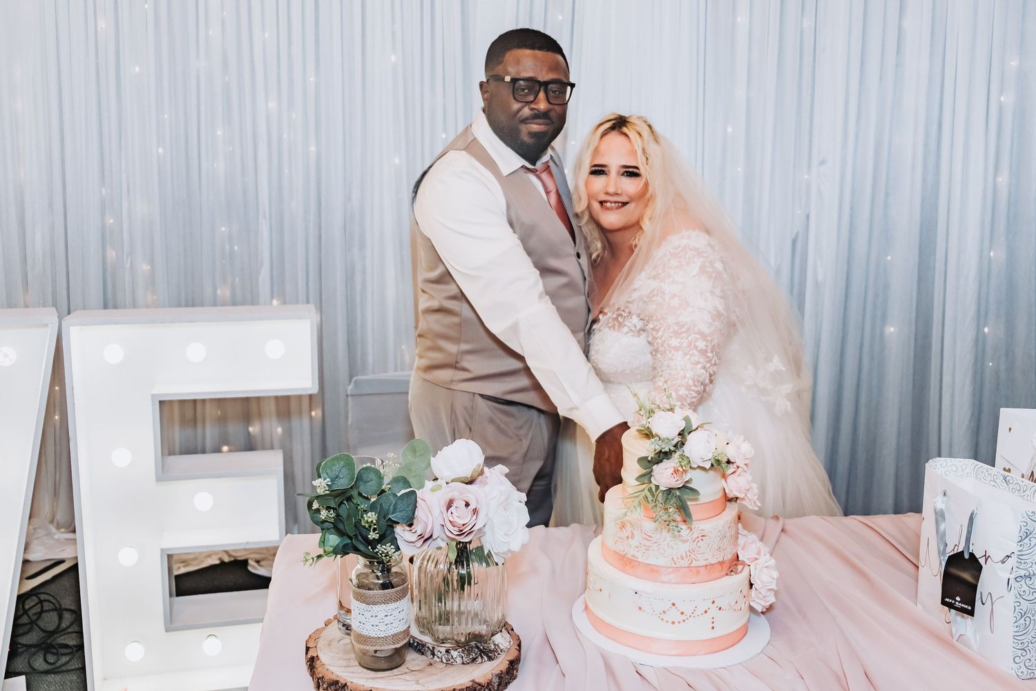 A couple smiles together at their wedding reception with a pink cake and decorative letter E on display.