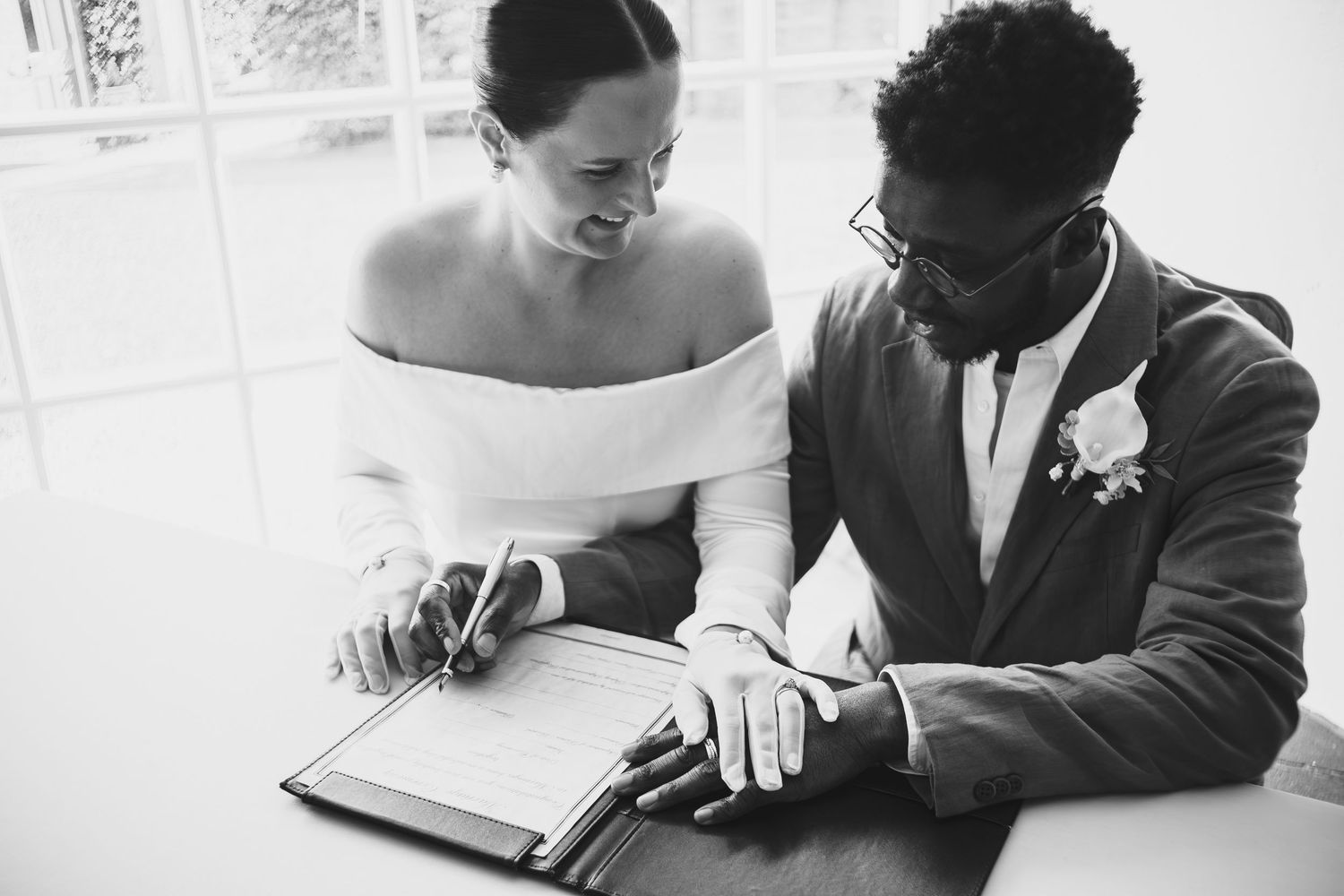 Black and white photo of a bride and groom signing their wedding registry document together at a table  at dunston hall estate