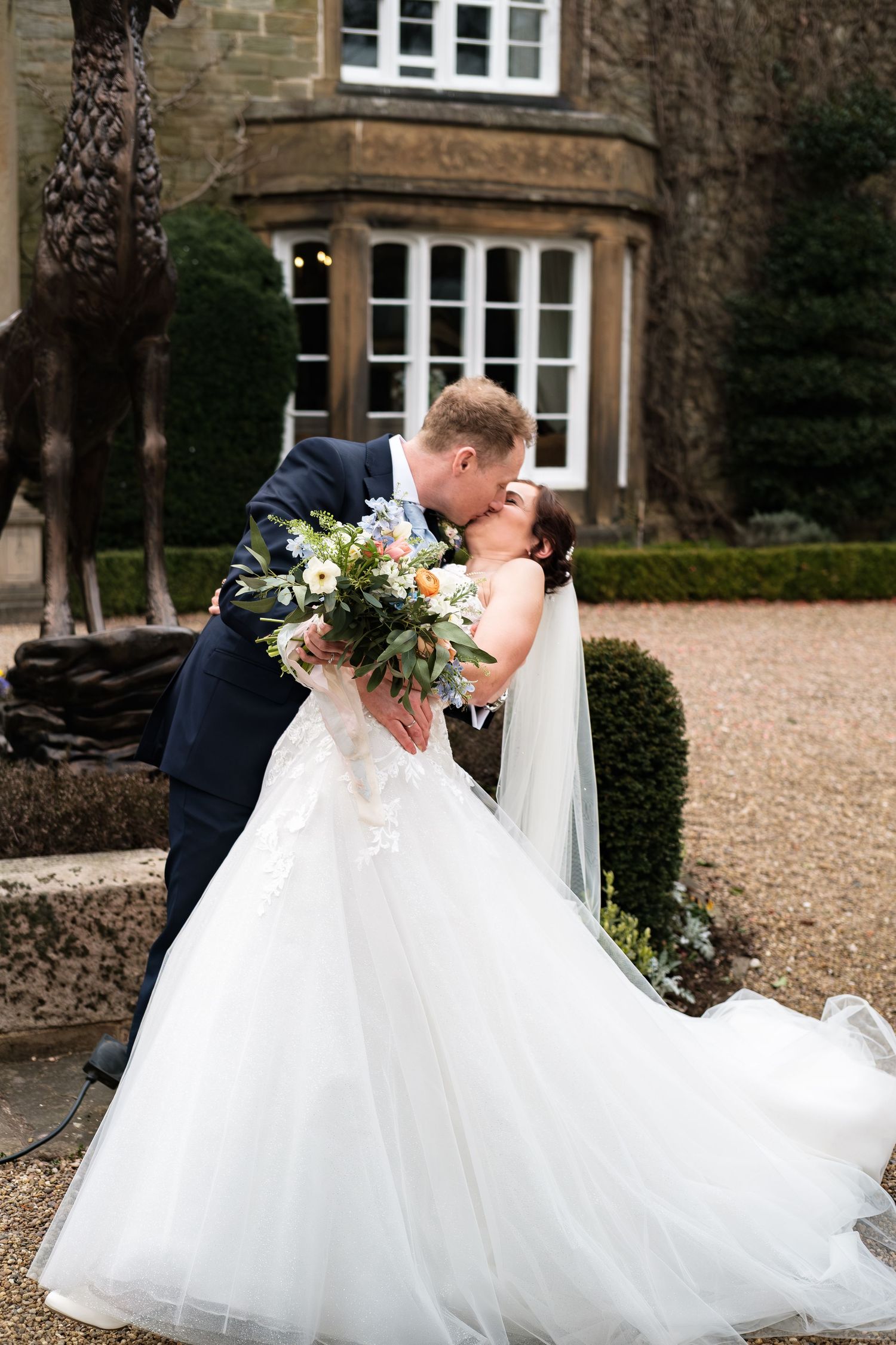 Bride and groom share romantic dip kiss outdoors holding bouquet in front of stone manor house.