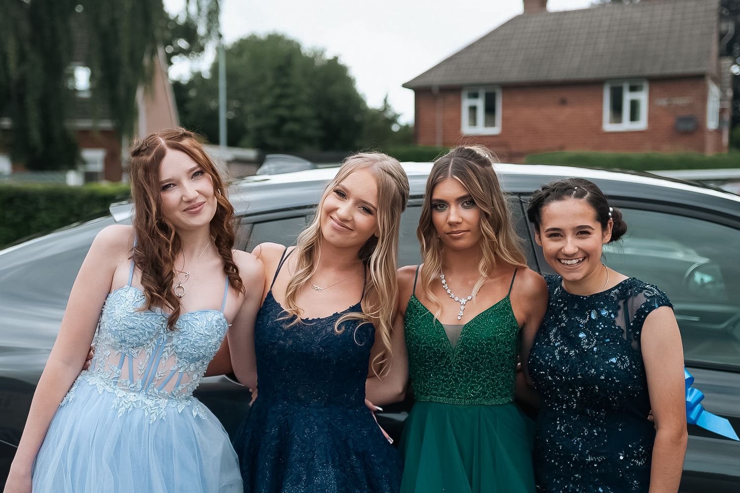 Group of four in formal dresses posing together outside near parked cars.
