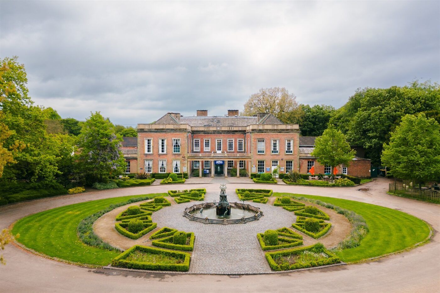 Elegant brick mansion with circular driveway featuring ornate gardens and fountain centerpiece surrounded by lush greenery.