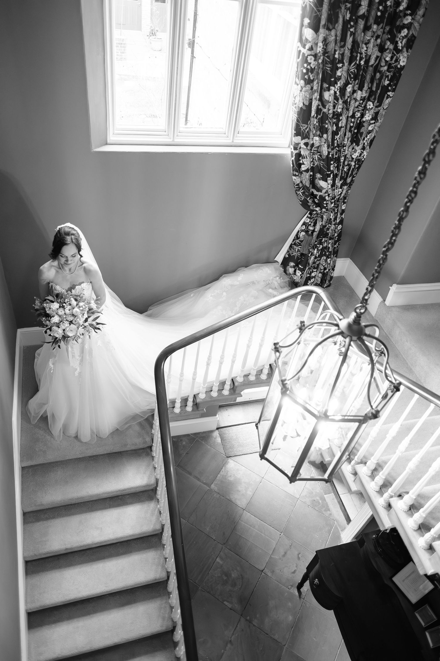 Bride in white gown holding bouquet descends elegant staircase with chandelier in black and white wedding photo.