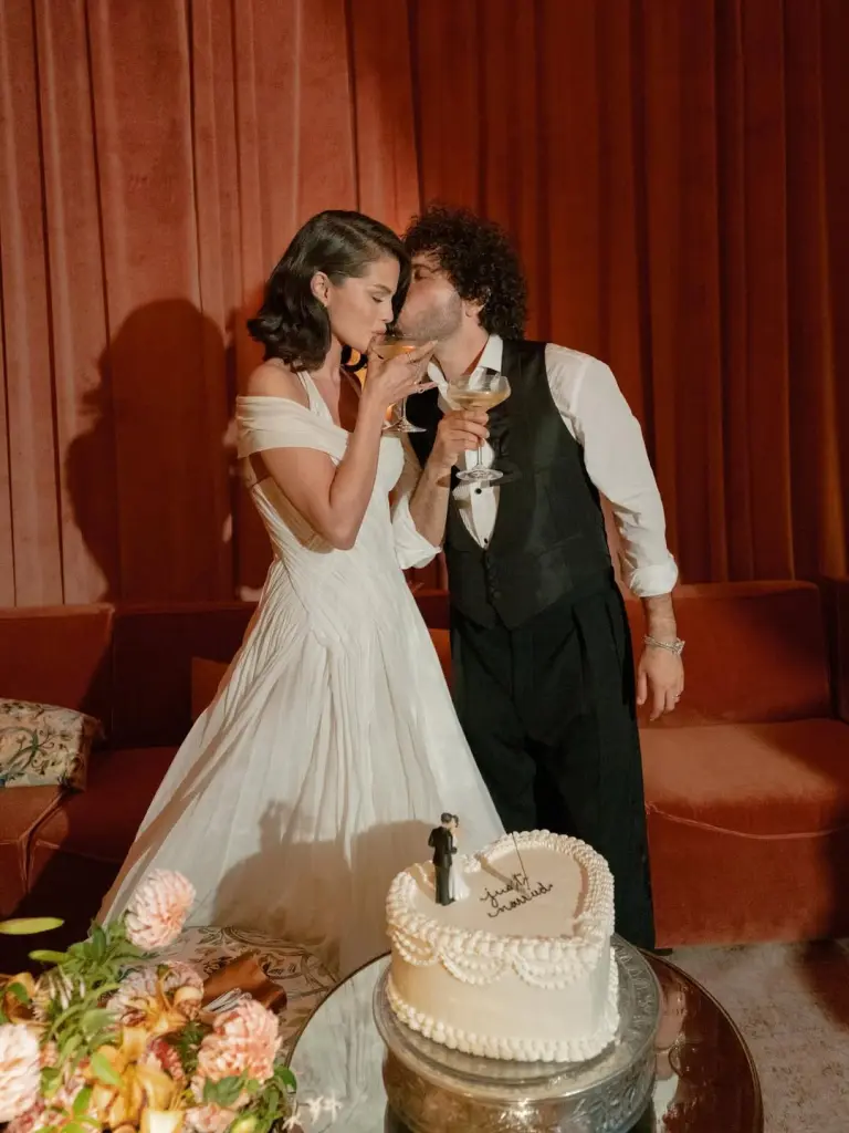 A couple shares a romantic kiss next to their wedding cake at their reception with warm red lighting in the background.