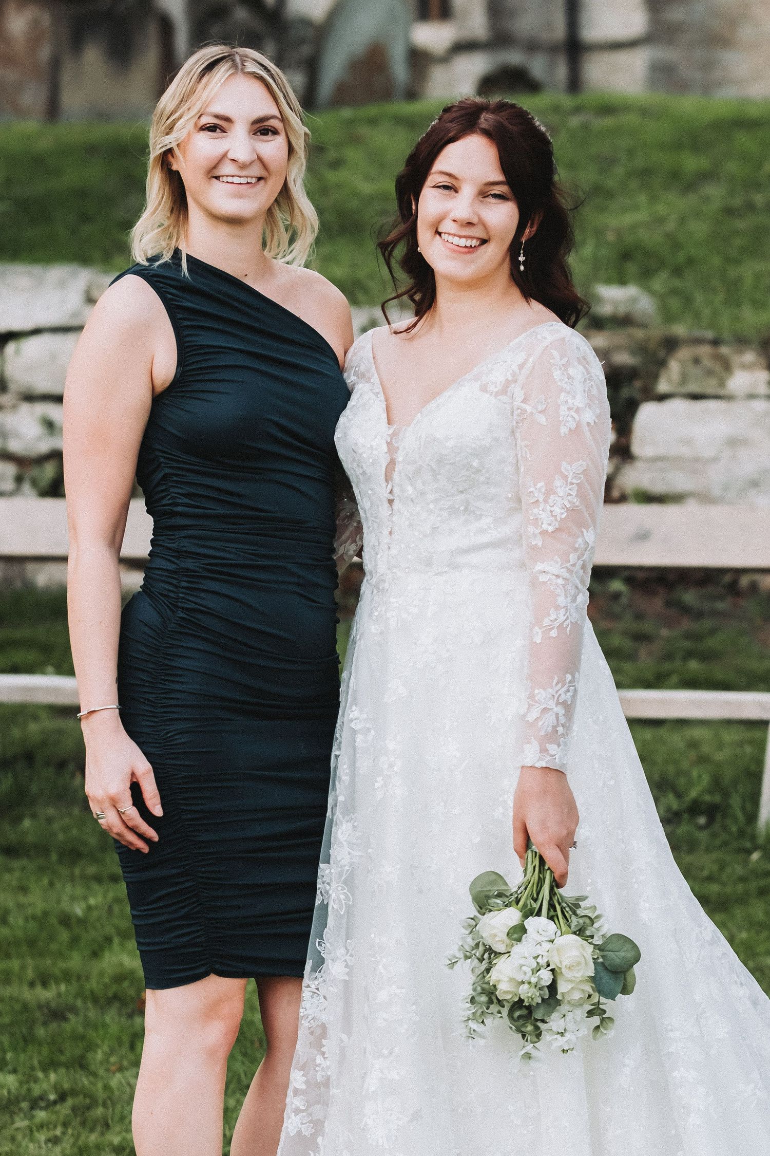 Bridesmaids in black dresses pose with bride wearing lace white gown and holding white bouquet outdoors.
