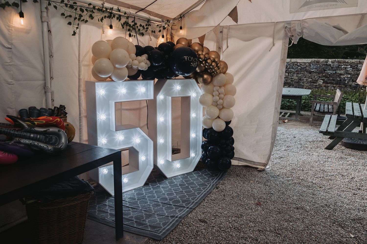 White wedding tent reception setup with twinkling lights, balloon decor and rustic wood details on a gravel surface.
