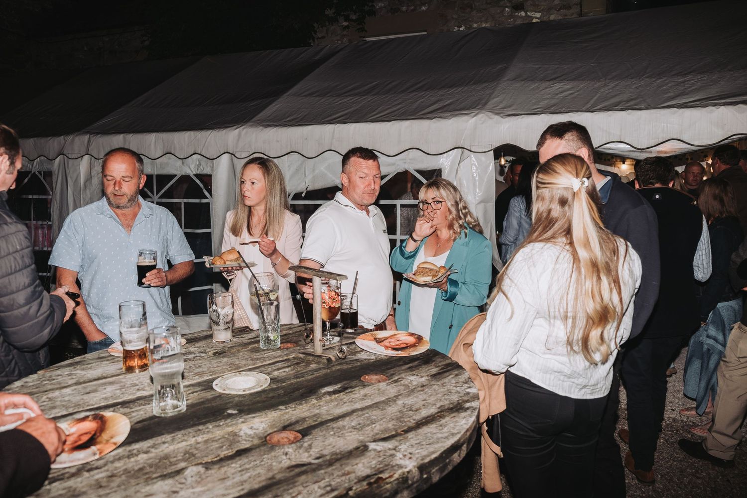 Large group of people gathered at a social event under a tent structure in evening lighting.