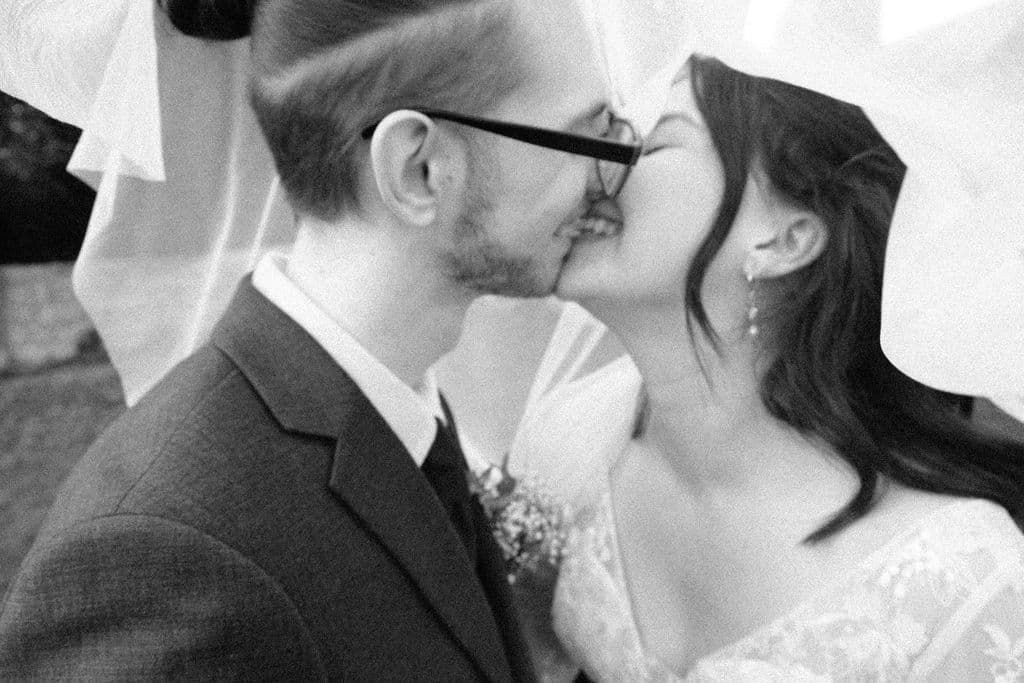 Black and white photo of a bride and groom sharing a kiss under a veil on their wedding day.