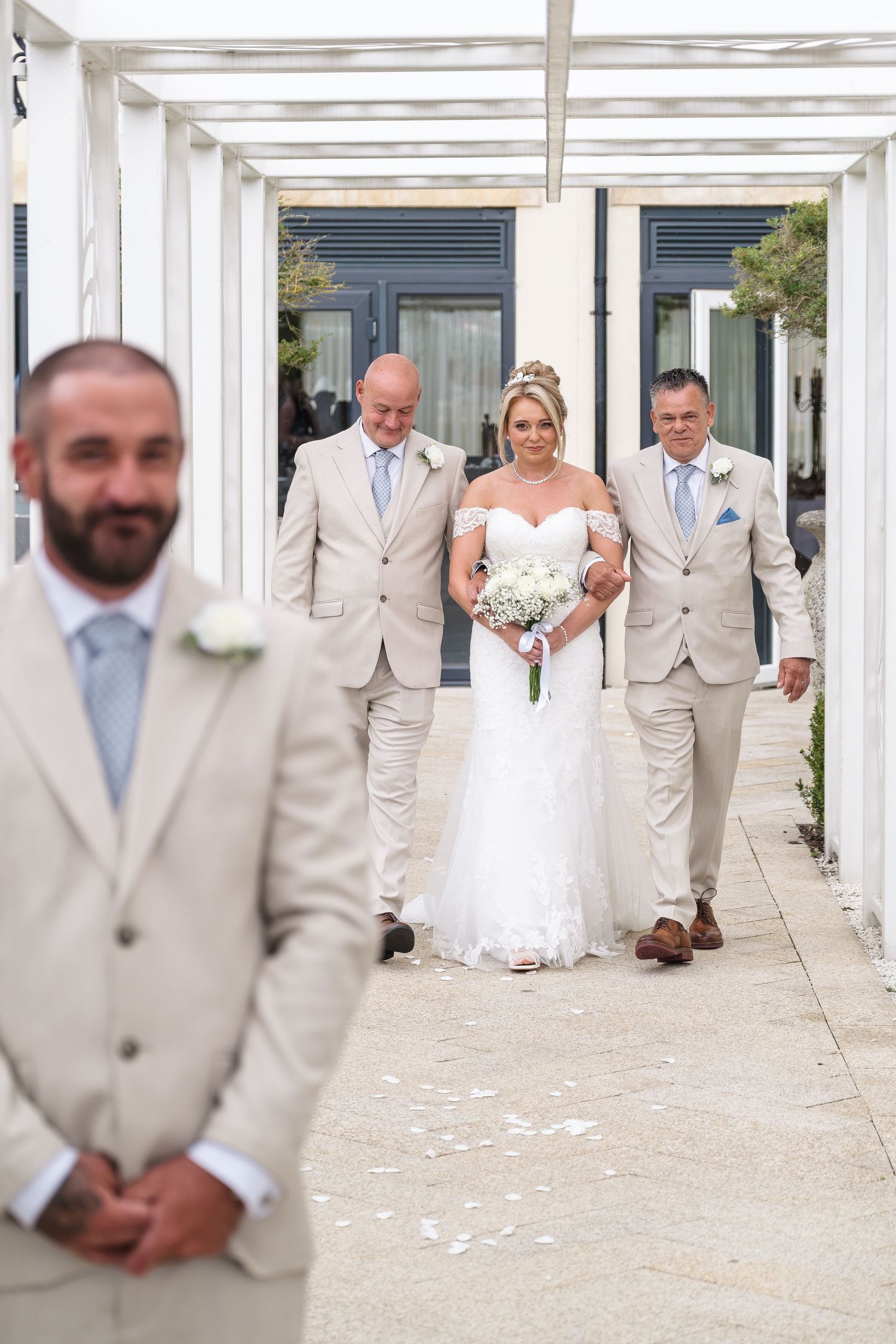 Bride in white gown walks down outdoor aisle with two groomsmen in beige suits, groom stands in foreground.