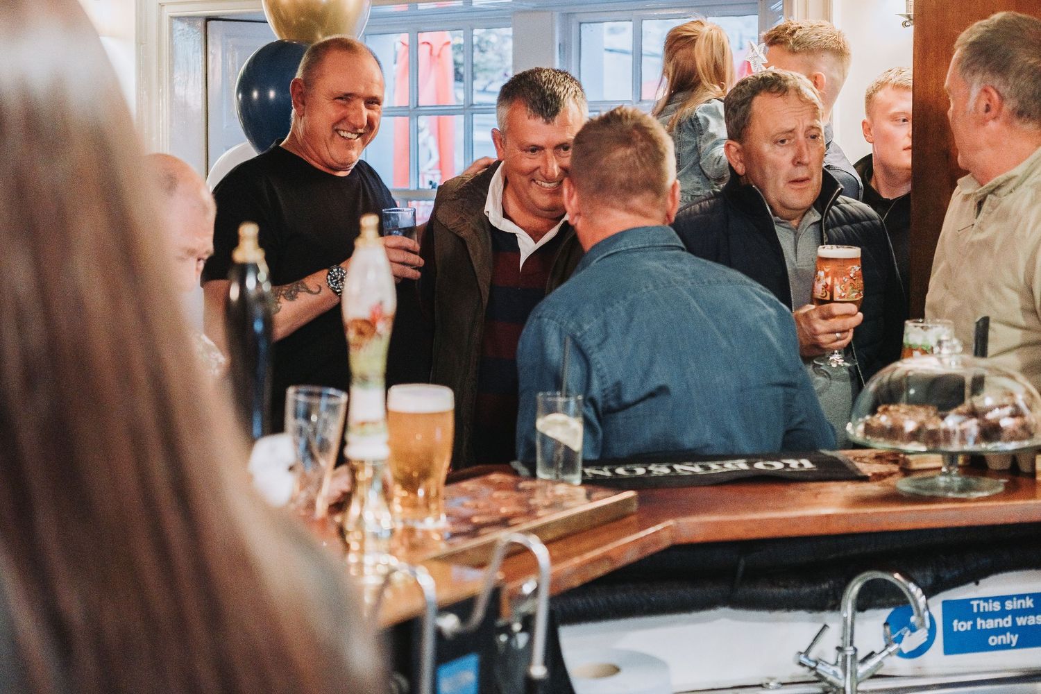 People enjoying drinks and conversation at a crowded bar with warm lighting and festive atmosphere.